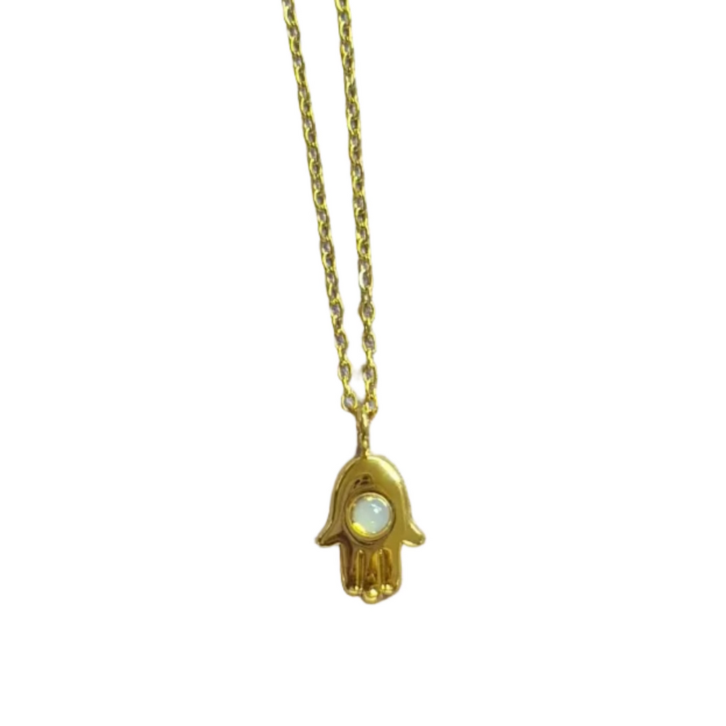 A gold hamsa hand pendant necklace with a small white inlay, shown on a light gray surface with a pink measuring ruler at the bottom for scale.